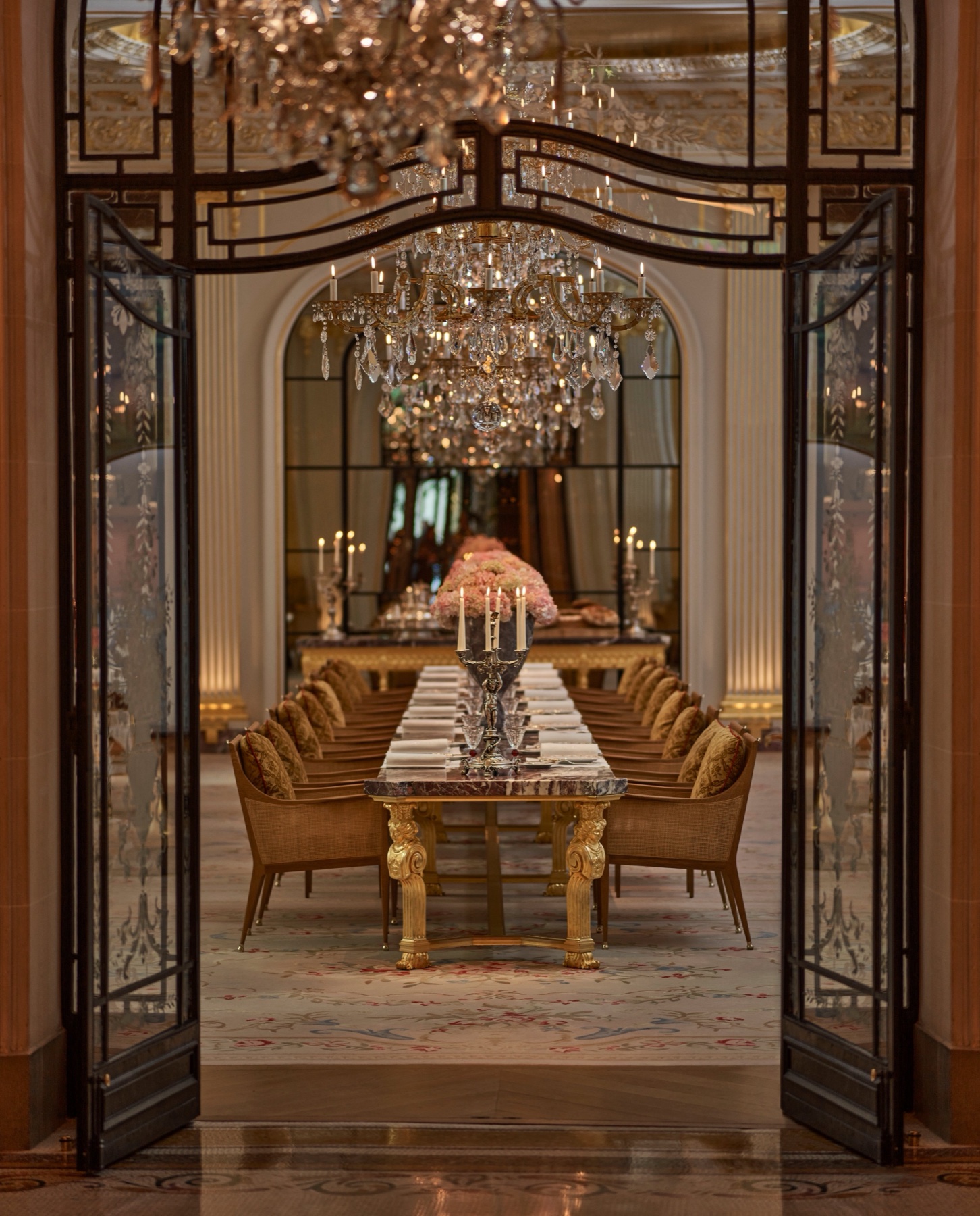 Vue à travers les portes en verre et fer forgé vers la longue table en marbre et les lustres de la salle gastronomique du Plaza Athénée