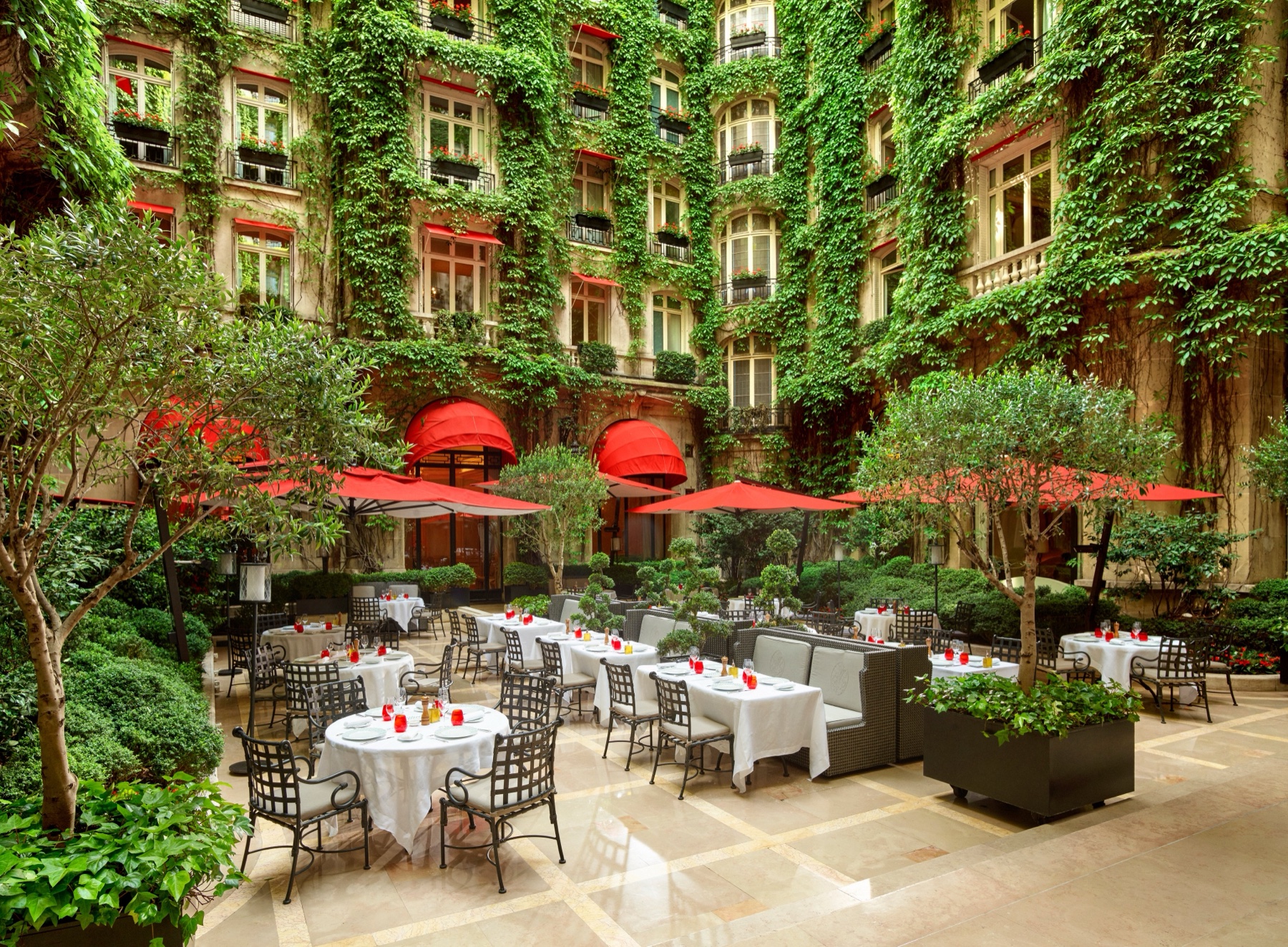 Terrasse de La Cour Jardin à l’Hôtel Plaza Athénée, entourée de façades couvertes de lierre, de stores rouges et de tables de jardin